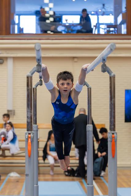 Focused young boy grips parallel bars mid-routine, arms raised, intense concentration on his face in a gymnasium setting.