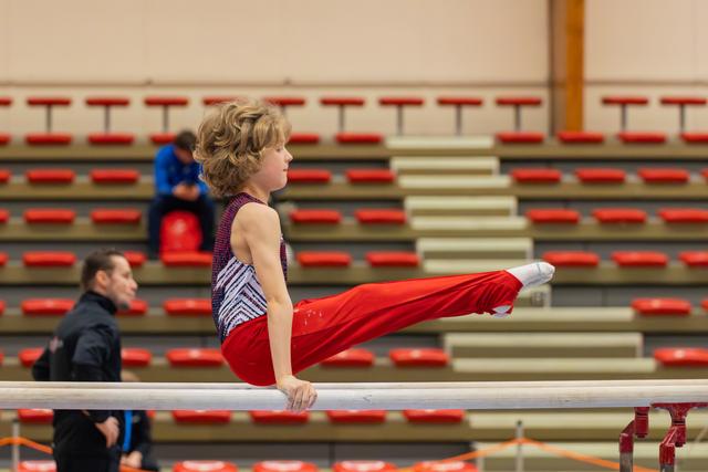 Young gymnast holds a precise L-sit on parallel bars, legs extended forward, focused expression in a nearly empty red-seated arena.