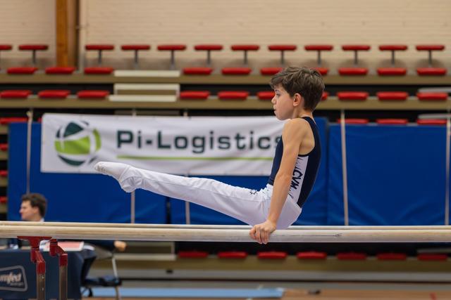 Focused boy holds a straddle-L on parallel bars, legs extended, concentration visible on his face in a gymnastics hall.