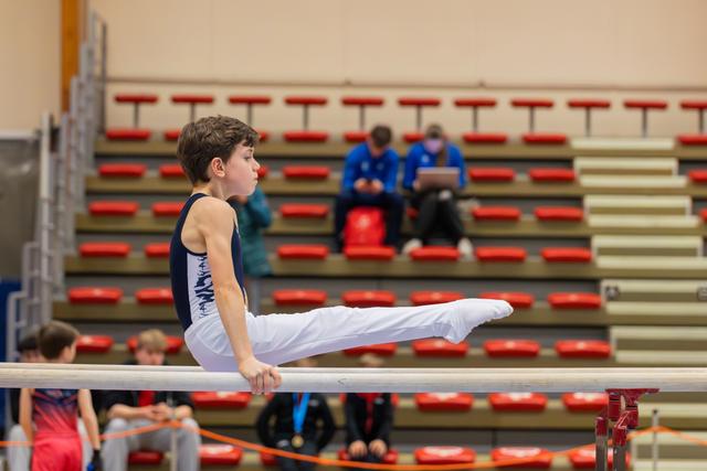 Focused young boy performs a straddle hold on parallel bars, legs extended, in a gymnasium with red bleacher seats.