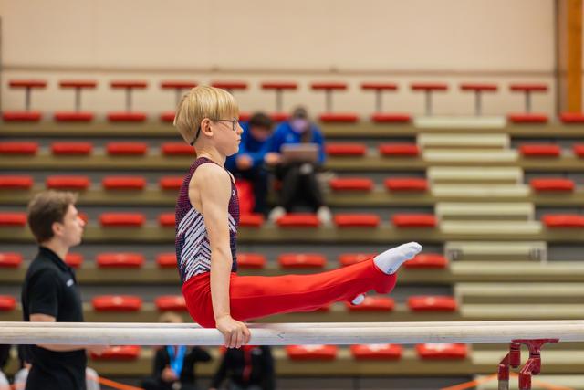 Blond boy with glasses performs a straddle hold on parallel bars, focused and composed, in a gymnastics hall.