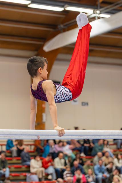 Young male gymnast holds an inverted pike position on parallel bars, legs raised high in red trousers, focused expression.