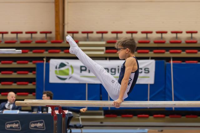Young boy in glasses performs a precise leg raise on parallel bars, showing intense focus during a gymnastics meet.