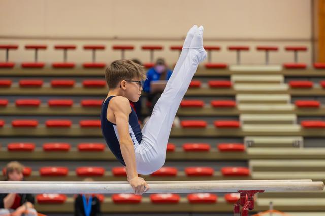 Young male gymnast in navy leotard executes a precise leg lift on parallel bars, wearing glasses, intense concentration.