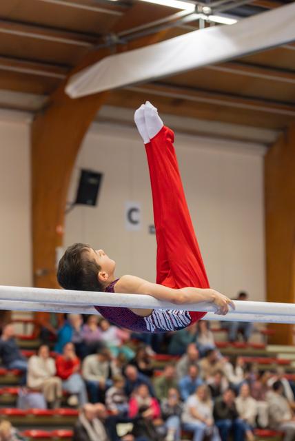 Young gymnast holds parallel bars with legs raised high, performing with focus and control before a seated crowd.