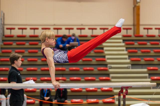Young gymnast with glasses holds a straddle position on parallel bars, legs raised high, focused expression in a gymnasium.