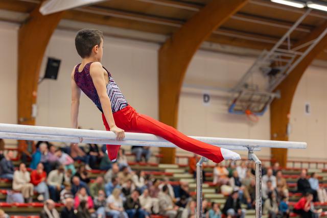 Young male gymnast holds a straddle position on parallel bars, focused and controlled, before a packed gymnasium crowd.