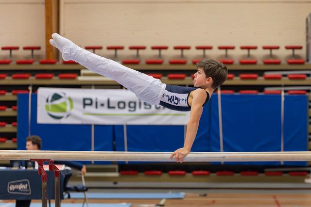 Young male gymnast holds a straddle position on parallel bars, body horizontal, focused expression in an indoor gymnasium.