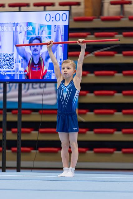 Young gymnast holds red stick overhead with focused expression, performing in blue leotard at Belgium gymnastics event.