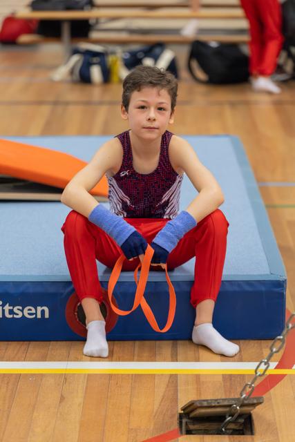 Young male gymnast sits on a blue mat holding orange straps, looking calm and focused between routines in a gym.