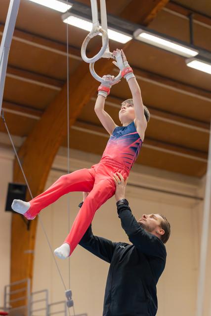 Young boy hangs focused on gymnastics rings while coach carefully spots him from below in an indoor gym.
