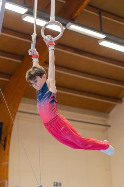 Young boy in red leotard swings on gymnastic rings with intense focus, body arched gracefully in a gym.