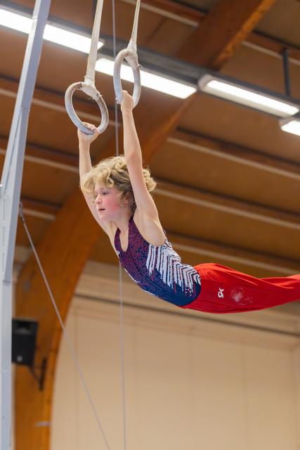 Young gymnast holds a horizontal position on still rings, focused expression, in a wood-beamed sports hall.
