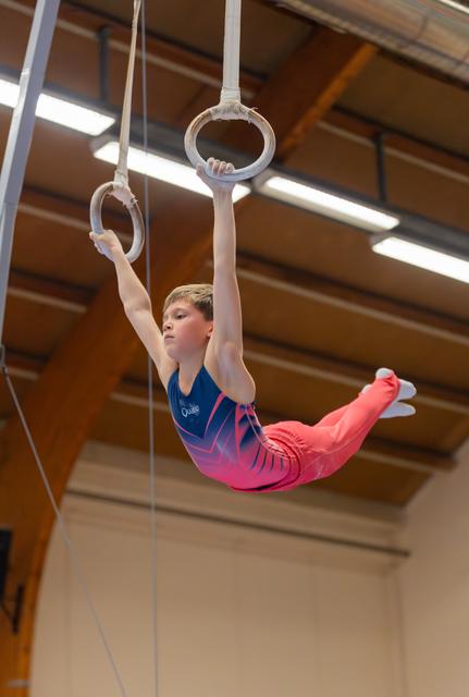 Young male gymnast holds gymnastics rings in a pike position, focused expression, wearing blue and pink leotard in an indoor gym.