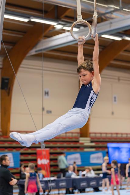 Young boy performs pike hang on gymnastics rings, legs extended parallel, focused expression in a busy sports hall.