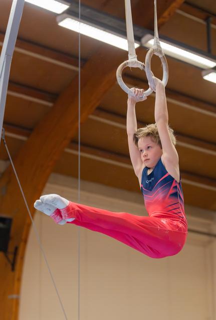 Young male gymnast holds gymnastics rings mid-air, legs in a wide split, wearing red and blue leotard, focused expression.