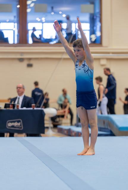 Young male gymnast raises both arms in a salute pose on the floor exercise mat, focused and composed at a gymnastics meet.