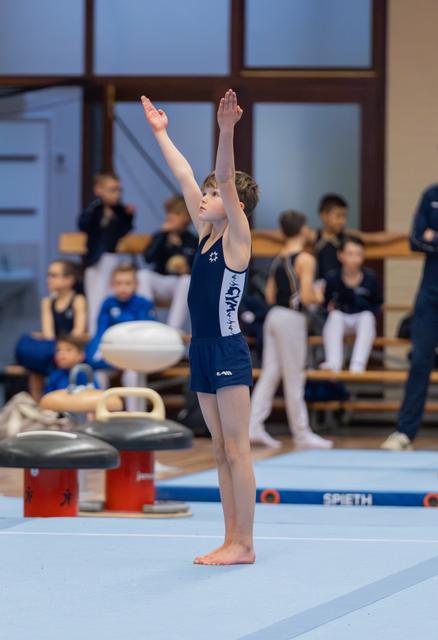 Young gymnast stands tall on the mat, arms raised in a proud salute, facing the judges in an indoor gymnastics hall.