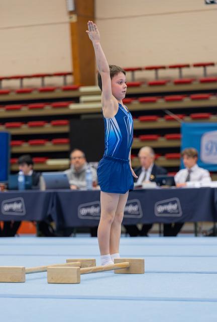 Young gymnast in blue leotard raises one arm in a confident salute on the floor exercise mat, judges seated behind.