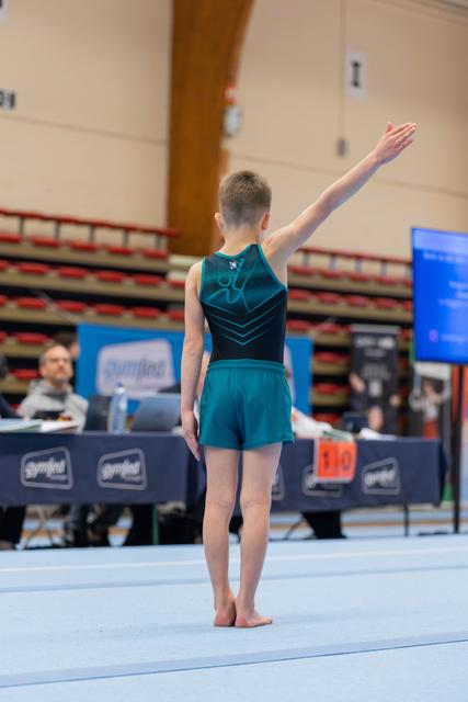 Young male gymnast stands barefoot on the floor mat, arm raised in a salute to the judges before his routine.