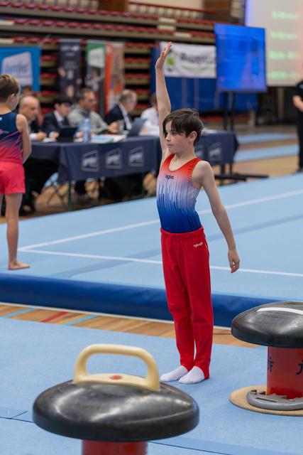 Young male gymnast raises one hand to salute judges before his routine, standing confidently on the blue floor mat.