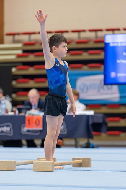 Young boy gymnast raises hand to salute judges, standing confidently on pommel horse blocks at indoor gymnastics meet.