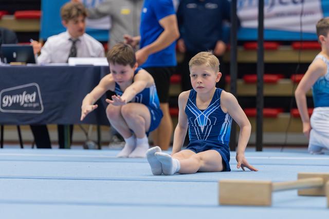 Young boy in blue leotard sits on the floor mat, composed and alert, during a gymnastics event judged by officials.