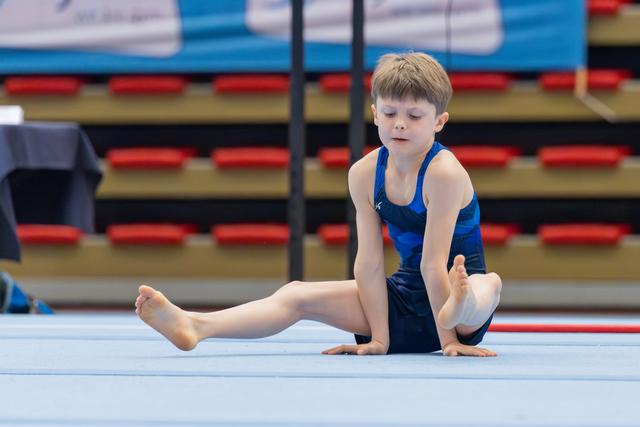 Young boy gymnast seated on floor mat, stretching with concentration, wearing blue leotard at indoor gymnastics event.