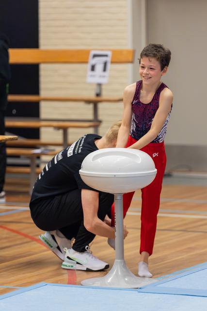 Smiling boy in gymnastics attire leans on mushroom apparatus while coach crouches to adjust equipment in a sports hall.