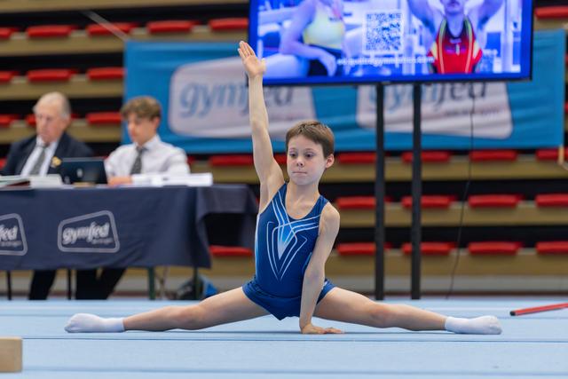 Young gymnast performs a split on the floor exercise mat, arm raised, focused expression, judges seated in background.