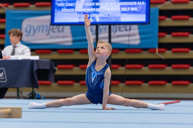 Blond child gymnast performs splits with arm raised high, focused expression, on blue floor at indoor gymnastics meet.