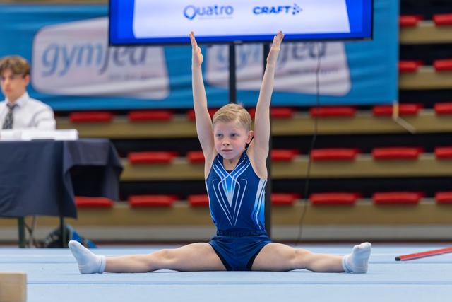 Young gymnast performs splits with arms raised high, focused expression, during a gymnastics meet on a blue floor.
