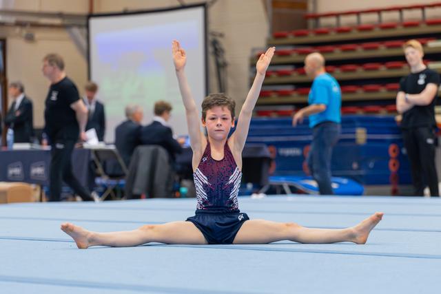 Young gymnast performs a split on the floor with arms raised high, focused expression during a gymnastics meet.