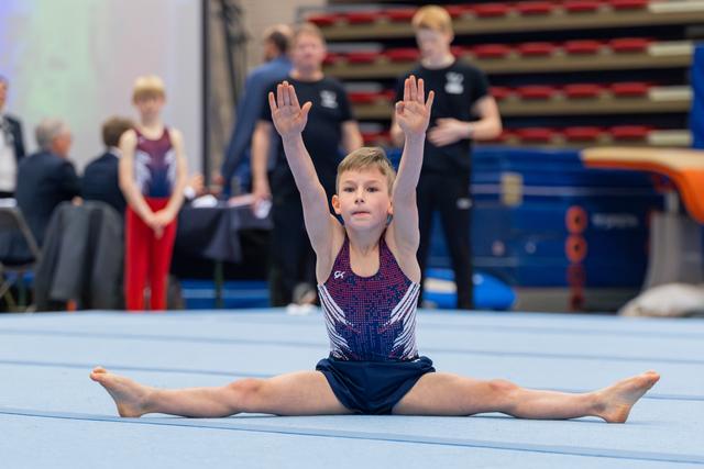 Young boy gymnast performs a split on the floor mat, arms raised high with focused expression during a gymnastics meet.
