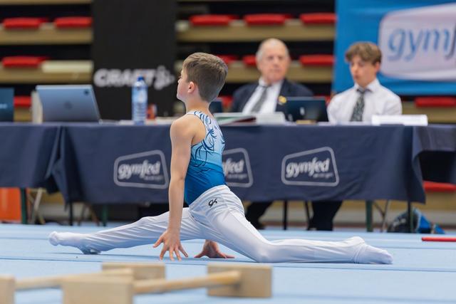 Young male gymnast performs a split on the floor, focused and poised, while judges observe from a table behind him.