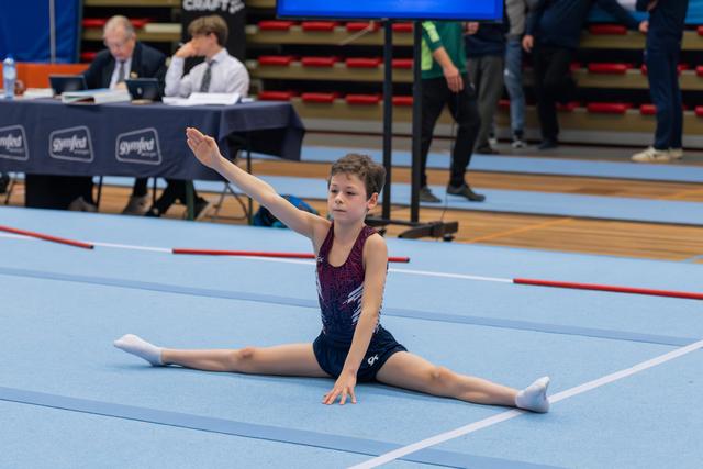 Young gymnast performs a straddle split on the floor exercise mat, arm raised gracefully, with judges seated at a table behind.