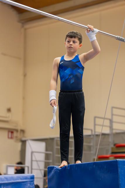 Young boy gymnast stands barefoot on a block, gripping the high bar with focused concentration, wearing blue leo and wrist wraps.