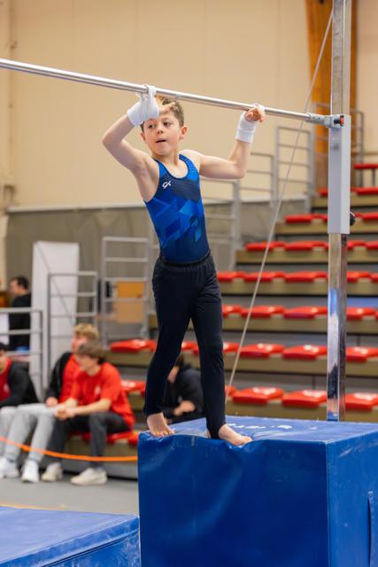 Young boy in blue leotard stands on mat gripping the high bar, focused expression, white wrist guards, indoor gym.