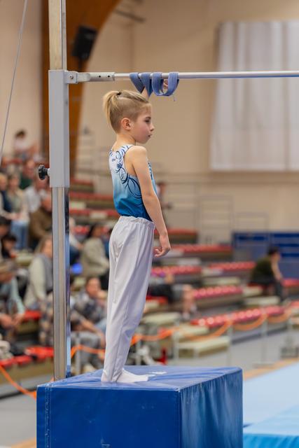 Young gymnast stands focused on a blue mat block, poised beneath the horizontal bar, awaiting their routine in a busy gymnasium.