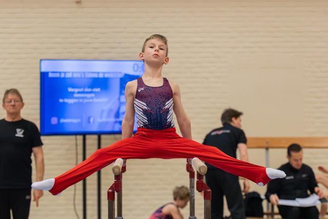 Young boy performs a straddle hold on parallel bars, legs split wide, with a focused, confident expression.