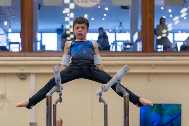 Young boy performs a straddle hold on parallel bars, legs spread wide, expression serious and concentrated in a gymnasium.