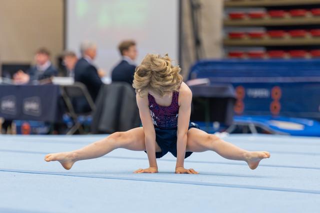 Young gymnast performs a straddle split on the blue floor mat, leaning forward with flowing blonde hair, judges visible behind.
