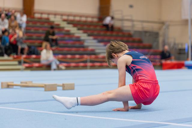 Young gymnast in red and navy leotard performs a split stretch on the blue floor mat, head bowed in focus.