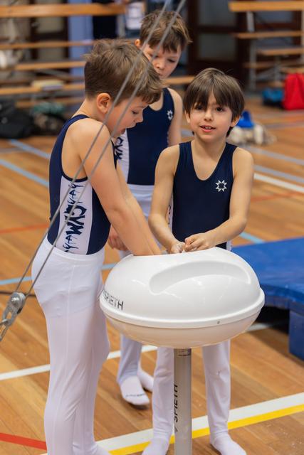 Three young male gymnasts in navy leotards apply chalk at a pommel horse, focused and preparing before their routine.