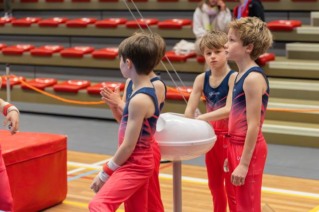 Three young male gymnasts in red and navy uniforms chalk their hands at a stand, focused and composed before their routine.
