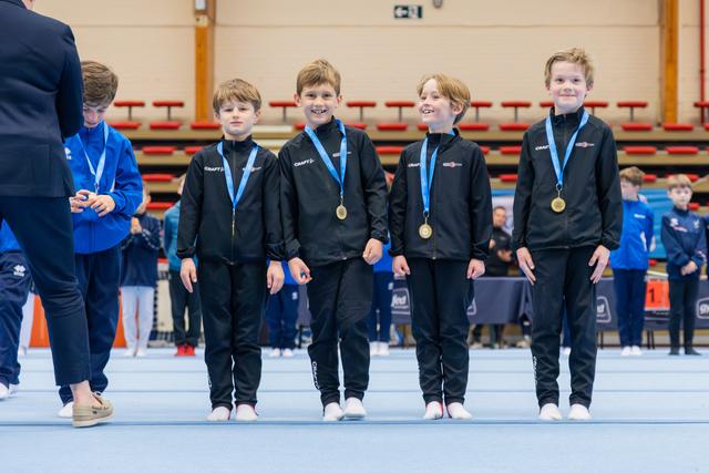 Four young boys in black tracksuits proudly display gold medals at a gymnastics competition, smiling on the competition floor.