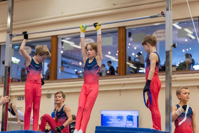 Three boys in red leotards hang and stand on gymnastics high bar, focused and determined, in a bright gymnasium.