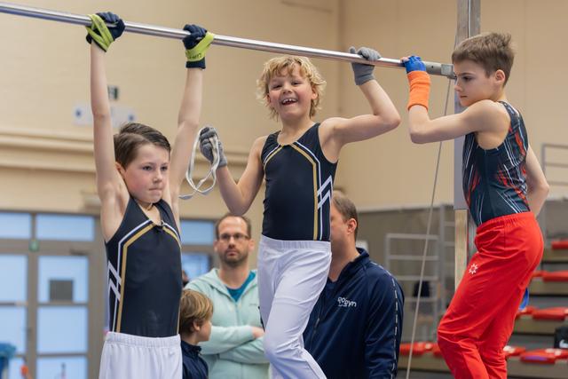 Three young male gymnasts hang from a high bar, the middle boy grinning broadly, watched by a coach in a gym.
