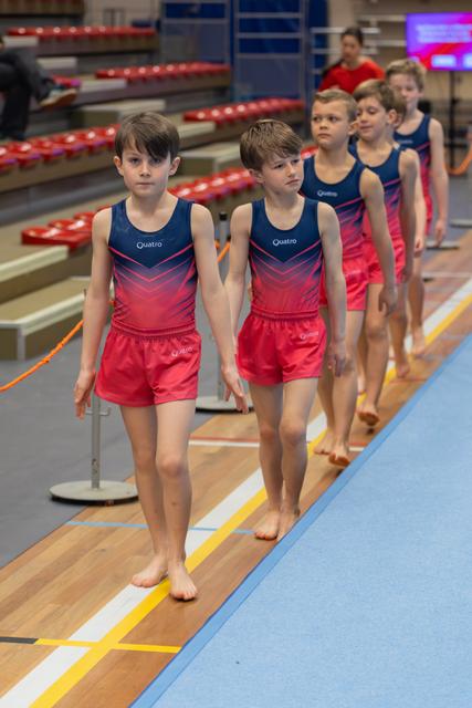 A line of young male gymnasts in matching Quatro uniforms walk barefoot across the floor, focused and composed.