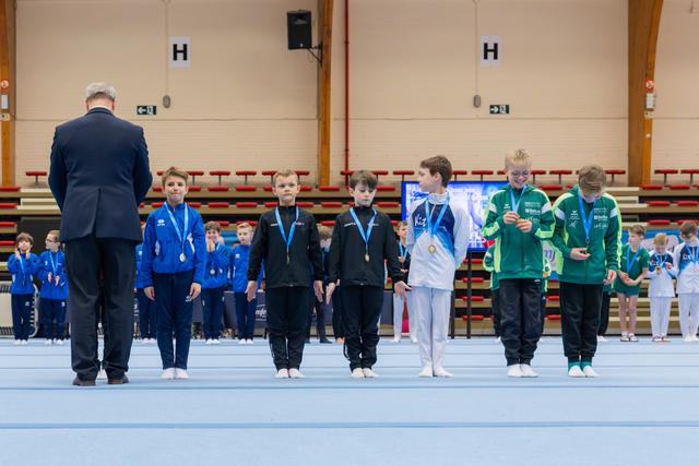 Young male gymnasts stand proudly on the gym floor receiving medals from an official during an indoor gymnastics competition.
